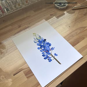 May include: A watercolor painting of a bluebonnet flower on white paper. The flower is blue with a white center and green stem.