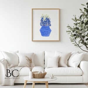May include: A watercolor painting of a blue vase with a white and blue pattern filled with blue and yellow flowers. The painting is framed in a light wood frame and is hanging on a white wall.