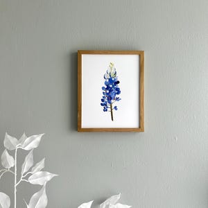 May include: A framed watercolor painting of a bluebonnet flower, a Texas state flower, with a white center and green stem. The painting is in a light brown frame.