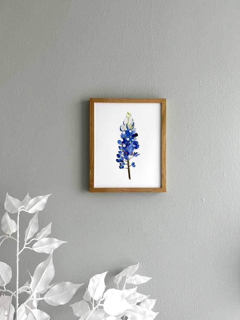 May include: A framed watercolor painting of a bluebonnet flower, a Texas state flower, with a white center and green stem. The painting is in a light brown frame.