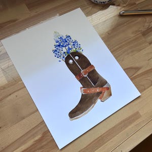 Puede incluir: Una bota vaquera de cuero marr&oacute;n con un arreglo de flores azules y blancas en la parte superior. La bota tiene una correa de cuero marr&oacute;n con una hebilla y un tac&oacute;n de cuero marr&oacute;n.