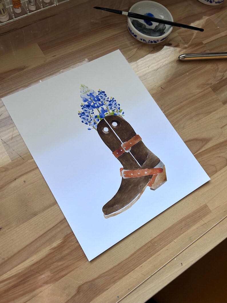 Puede incluir: Una bota vaquera de cuero marr&oacute;n con un arreglo de flores azules y blancas en la parte superior. La bota tiene una correa de cuero marr&oacute;n con una hebilla y un tac&oacute;n de cuero marr&oacute;n.