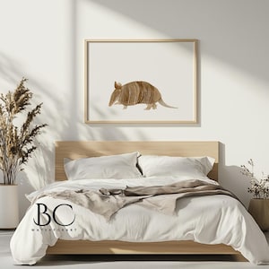 May include: A watercolor painting of an armadillo on a white background. The armadillo is brown and has a textured shell. The painting is framed in a light wood frame and is hanging on a white wall above a bed with white bedding.