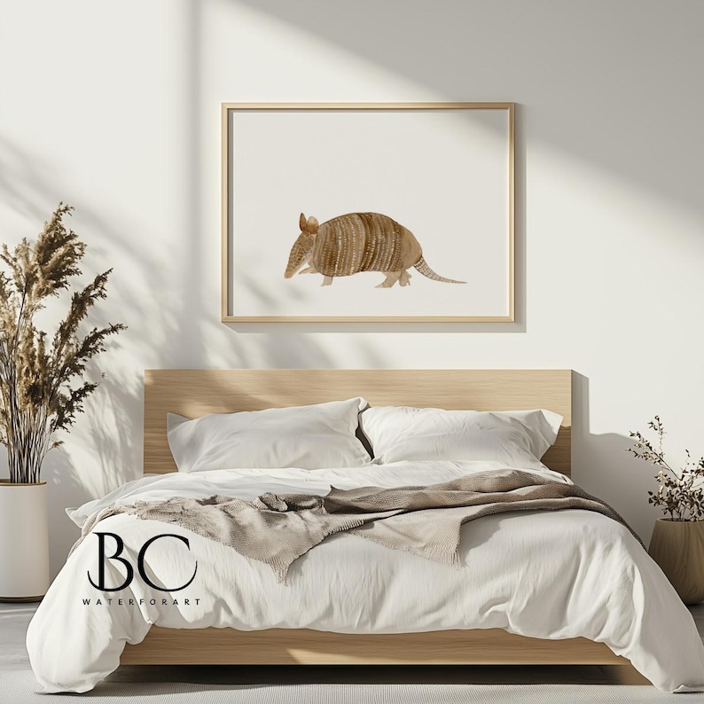 May include: A watercolor painting of an armadillo on a white background. The armadillo is brown and has a textured shell. The painting is framed in a light wood frame and is hanging on a white wall above a bed with white bedding.