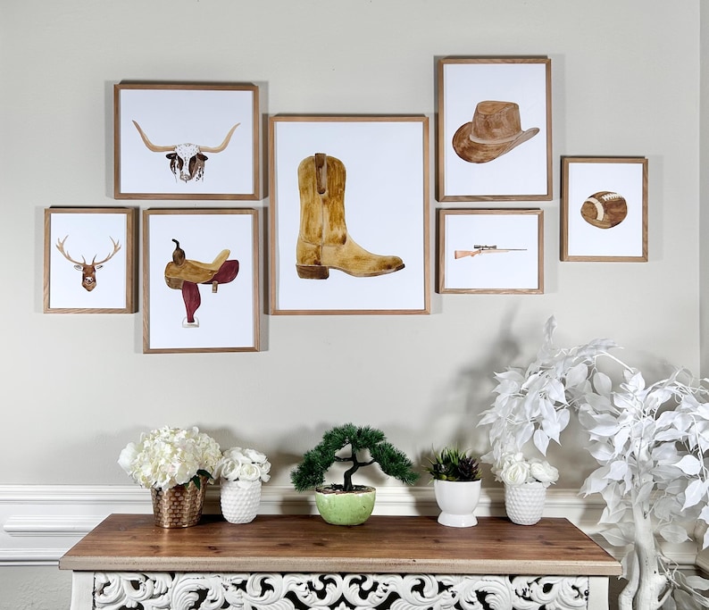 May include: A set of framed watercolor prints with a western theme. The prints feature a longhorn skull, a cowboy boot, a saddle, a deer head, a cowboy hat, a rifle, and a football.