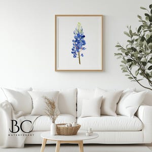 May include: A watercolor painting of a bluebonnet flower, a Texas state flower, in a wooden frame. The painting is hanging on a white wall above a white couch with throw pillows.