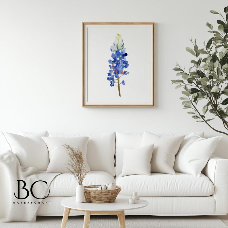 May include: A watercolor painting of a bluebonnet flower, a Texas state flower, in a wooden frame. The painting is hanging on a white wall above a white couch with throw pillows.