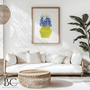 May include: A watercolor painting of a bouquet of bluebonnet flowers in a yellow woven vase. The painting is framed and hanging on a white wall in a living room setting.
