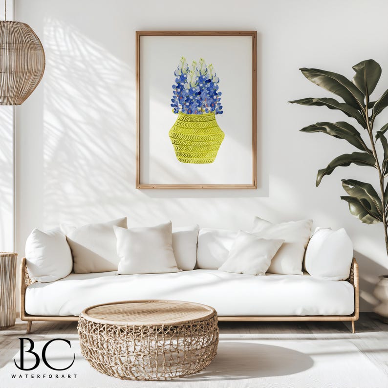 May include: A watercolor painting of a bouquet of bluebonnet flowers in a yellow woven vase. The painting is framed and hanging on a white wall in a living room setting.