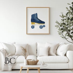 May include: A framed watercolor print featuring a blue roller skate with gold accents. The artwork is set against a white background and framed in a light wood. The print is displayed above a white sofa in a living room setting.