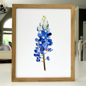 May include: A framed watercolor painting of a bluebonnet flower. The flower is depicted in shades of blue, green, and white, with a yellow center. The painting is set against a white background.