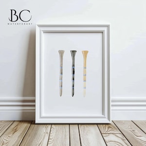 May include: A framed watercolor painting of three golf tees in different colors: beige, gray, and brown. The tees have blue stripes.