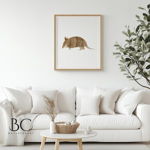 May include: A watercolor painting of an armadillo in a light brown frame. The armadillo is facing left and has a light brown shell with dark brown stripes. The background is white.