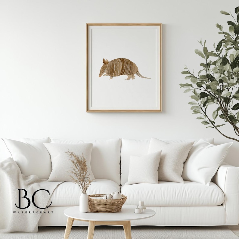 May include: A watercolor painting of an armadillo in a light brown frame. The armadillo is facing left and has a light brown shell with dark brown stripes. The background is white.