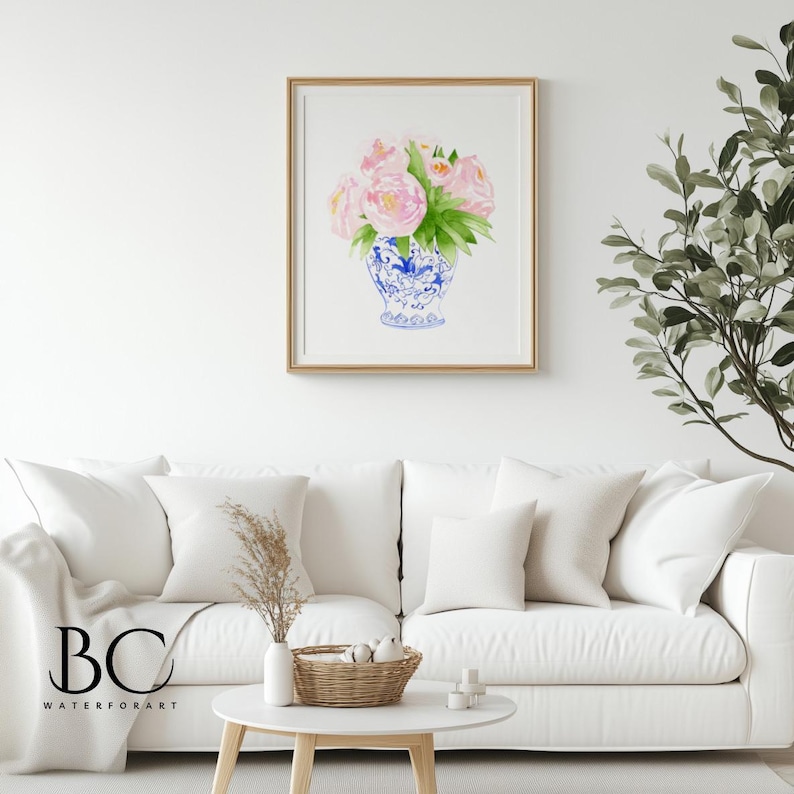 Puede incluir: Pintura a la acuarela de rosas rosas y hojas verdes en un jarr&oacute;n azul y blanco. La obra de arte est&aacute; enmarcada en un marco dorado y cuelga de una pared blanca sobre un sof&aacute; blanco. La obra de arte est&aacute; etiquetada "BC WATERFORART".
