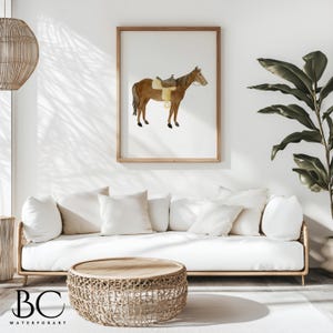 May include: A watercolor painting of a brown horse with a saddle on its back. The horse is standing on a white background. The painting is framed in a light wood frame and is hanging on a white wall.