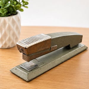 May include: A vintage Bostitch stapler with a matte olive green metal body and a textured silver-toned top plate. The stapler features a rectangular base, a hinged mechanism, and the brand name Bostitch embossed on the side of the metal casing.