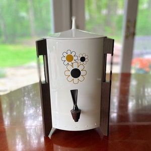 Puede incluir: Cafetera blanca con un soporte de madera marrón y un diseño de margarita blanco y negro. La cafetera tiene un pico negro y una tapa blanca.