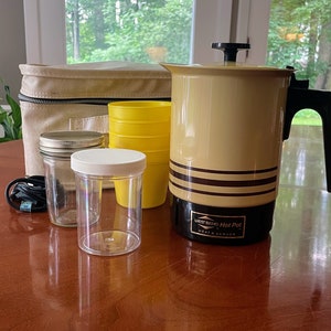 May include: A vintage West Bend Hot Pot with a beige and brown striped exterior. The hot pot is on a wooden table with a brown canvas bag, four yellow plastic cups, and two glass jars.