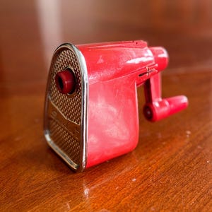 May include: A vintage red pencil sharpener with a silver-colored trim. The sharpener has a crank handle and a small opening for the pencil. The front of the sharpener has a textured, patterned design.