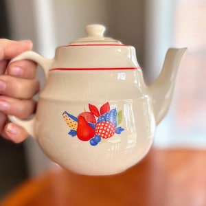 May include: Cream-colored ceramic teapot with a red band and fruit design. The teapot has a curved spout, a handle, and a lid with a small knob. The fruit design includes red, blue, and yellow elements.
