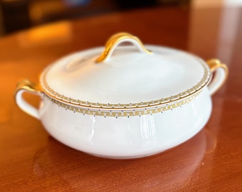 May include: White lidded serving bowl with gold accents. The bowl has two handles and a gold handle on the lid. A gold geometric pattern decorates the rim of the bowl. The bowl is on a wooden surface.