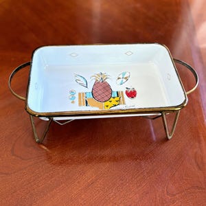 Könnte beinhalten: Ein rechteckiges Serviertablett aus weißem Emaille mit goldenem Rand und Griffen, das ein farbenfrohes Ananas- und Fruchtdesign aufweist. Das Tablett steht auf einem goldenen Metallständer. Das Design umfasst eine Ananas, eine Erdbeere und andere tropische Elemente.