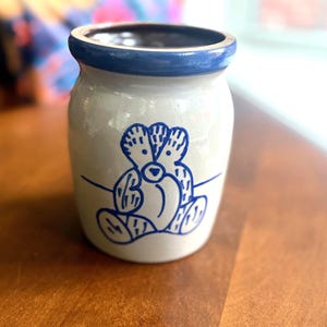 May include: A cylindrical stoneware crock with a light grey finish and a dark blue rim. The front features a hand-painted blue line drawing of a seated bear with textured fur details, centred on the body of the vessel with a horizontal line.