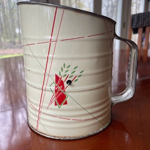 May include: White metal flour sifter with a red and green geometric design. The sifter has a handle and a lid.