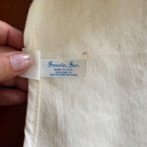 K&ouml;nnte beinhalten: Nahaufnahme eines cremefarbenen Stoffes mit einem Etikett mit der Aufschrift "Innovo, Inc. MADE IN U.S.A. STAFFORD, TX (SIEHE R&Uuml;CKSEITE ZUR PFLEGE)". Der Stoff hat eine subtile Webstruktur.