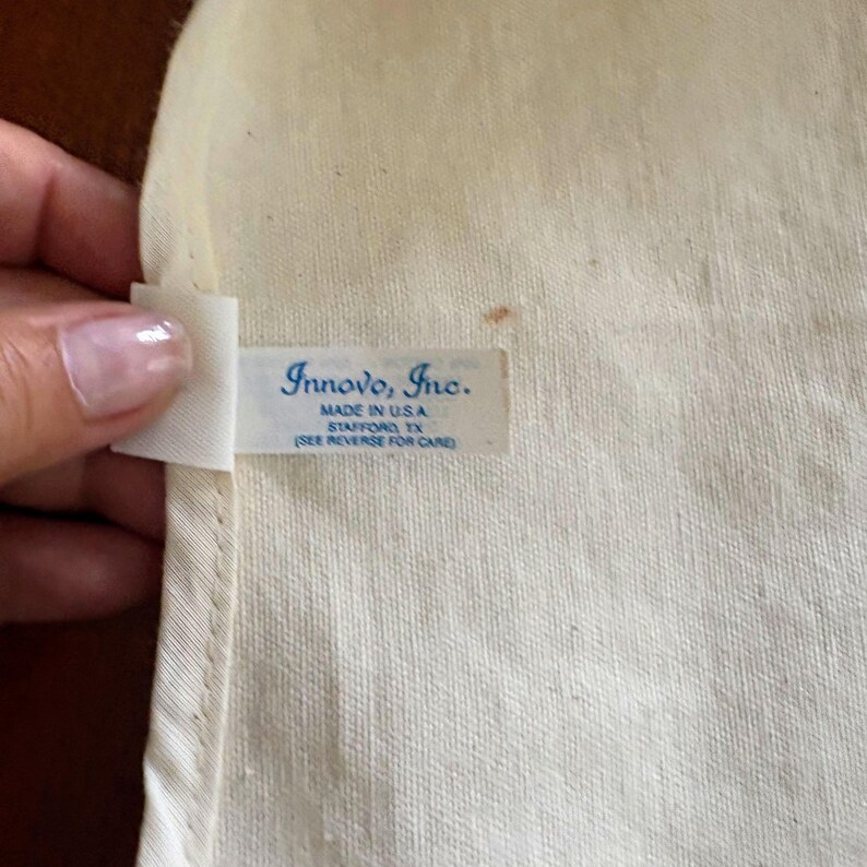 K&ouml;nnte beinhalten: Nahaufnahme eines cremefarbenen Stoffes mit einem Etikett mit der Aufschrift "Innovo, Inc. MADE IN U.S.A. STAFFORD, TX (SIEHE R&Uuml;CKSEITE ZUR PFLEGE)". Der Stoff hat eine subtile Webstruktur.