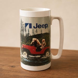 Op de afbeelding: Een witte cilindervormige mok met een groot handvat, voorzien van een afbeelding van een rode Jeep die door water rijdt. Het ontwerp toont een beer achter het stuur, een passagier, een bos op de achtergrond en het blauwe Jeep-logo bovenaan.