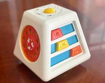 Puede incluir: Un juguete vintage en forma de pirámide de color blanco roto con un dial giratorio rojo y bloques deslizantes de colores. El juguete tiene un botón amarillo en la parte superior, diseñado para el desarrollo de la primera infancia.