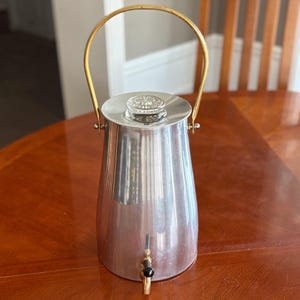 Può includere: Un server per bevande vintage color argento con manico color oro. Il server ha un coperchio in vetro trasparente e un rubinetto con manico nero. Il server viene probabilmente utilizzato per servire bevande fredde.