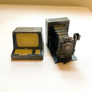 May include: Two vintage metal pencil sharpeners, one shaped like a computer and the other shaped like a camera. Both are silver in color.