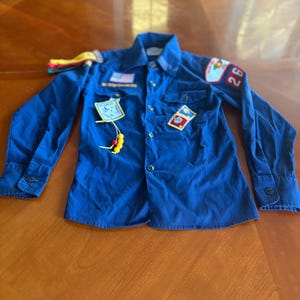 Könnte beinhalten: Ein dunkelblaues Hemd mit Knöpfen, verziert mit mehreren Aufnähern und Insignien. Das Hemd hat lange Ärmel und einen Kragen. Zu den Aufnähern gehören eine US-Flagge, die Zahl 26 und andere Embleme. Ein gelb-rotes Perlenband hängt am Hemd.