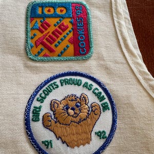 K&ouml;nnte beinhalten: Zwei gestickte Aufn&auml;her auf cremefarbenem Stoff. Einer zeigt "100" und "COOKIES 92" mit einem farbenfrohen geometrischen Design. Der andere Aufn&auml;her zeigt "GIRL SCOUTS PROUD AS CAN BE" mit einer B&auml;renillustration und den Zahlen "91" und "92".