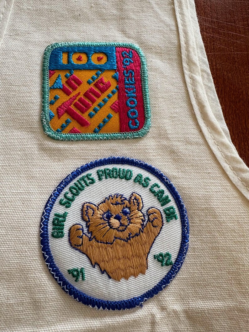 K&ouml;nnte beinhalten: Zwei gestickte Aufn&auml;her auf cremefarbenem Stoff. Einer zeigt "100" und "COOKIES 92" mit einem farbenfrohen geometrischen Design. Der andere Aufn&auml;her zeigt "GIRL SCOUTS PROUD AS CAN BE" mit einer B&auml;renillustration und den Zahlen "91" und "92".