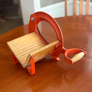 May include: Vintage red and wood bread slicer. The slicer has a wooden base and a red metal frame with a handle. The blade is not visible. The slicer is sitting on a wooden table.