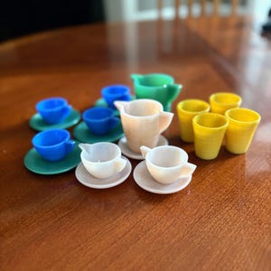 May include: A collection of miniature tea party dishes in various colors. The set includes blue cups and green saucers, yellow tumblers, and cream-colored teacups with saucers. A green and cream-colored creamer is also included.