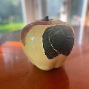 May include: A ceramic apple-shaped jar with a lid. The jar is painted with shades of yellow, brown, red, and green. The jar has a rustic, vintage look.