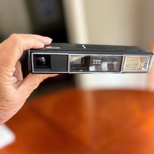 Puede incluir: Una cámara negra ITT Magicflash Teleflash con un borde plateado se sostiene en la mano. La cámara tiene un flash y una lente. El texto "ITT MAGICFLASH TELEFLASH CAMERA" está impreso en la parte delantera.