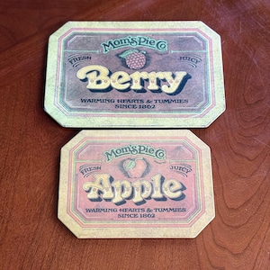 Peut inclure: Deux moules à tarte de style vintage avec le texte "Mom's Pie Co." et les mots "Berry" et "Apple" dans une police de caractères stylisée. Les moules sont jaunes, rouges et verts avec un fond brun. Le texte "Warming Hearts & Tummies Since 1862" est imprimé sur les deux moules.
