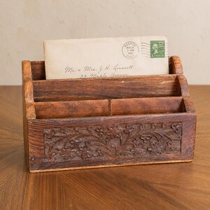 May include: A dark brown wooden letter holder with three tiered compartments and a carved floral pattern on the front panel. An off-white envelope is tucked into the back slot, featuring a Boston postmark dated May 18, 1921, and a green stamp.