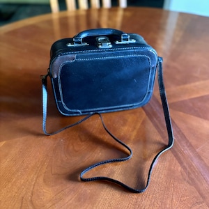Può includere: Una piccola borsa in pelle nera con manico e una tracolla lunga e sottile. La borsa ha una forma rettangolare con un pannello in camoscio sul davanti e dettagli in pelle marrone. La borsa è su un tavolo di legno.
