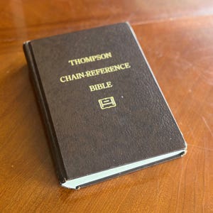 Könnte beinhalten: Eine braune, ledergebundene Bibel mit goldener Schrift. Der Text auf dem Einband lautet "THOMPSON CHAIN-REFERENCE BIBLE". Unter dem Wort "BIBLE" befindet sich ein kleines goldenes Emblem. Das Buch ist geschlossen und liegt auf einer Holzoberfläche.
