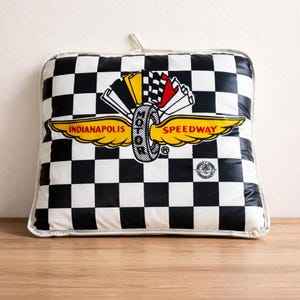 May include: A square pillow with a black and white checkered pattern. The pillow features a logo with the words "Indianapolis Speedway" in yellow, a tire graphic, and a checkered flag design.