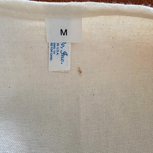 K&ouml;nnte beinhalten: Nahaufnahme eines cremefarbenen Stoffes mit einem wei&szlig;en Etikett. Das Etikett zeigt den Buchstaben "M", den Text "Jo, Inc." und den Standort "ORD, TX". Der Stoff hat ein leicht strukturiertes Aussehen.