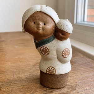 Może przedstawiać: Ceramiczna figurka przedstawiająca osobę niosącą na plecach mniejszą postać. Przedmiot posiada nakrapianą białą glazurę w górnych partiach, ciemnobrązową podstawę, ciemnoniebieski kołnierzyk oraz brązowe motywy kwiatowe na białym ubraniu.