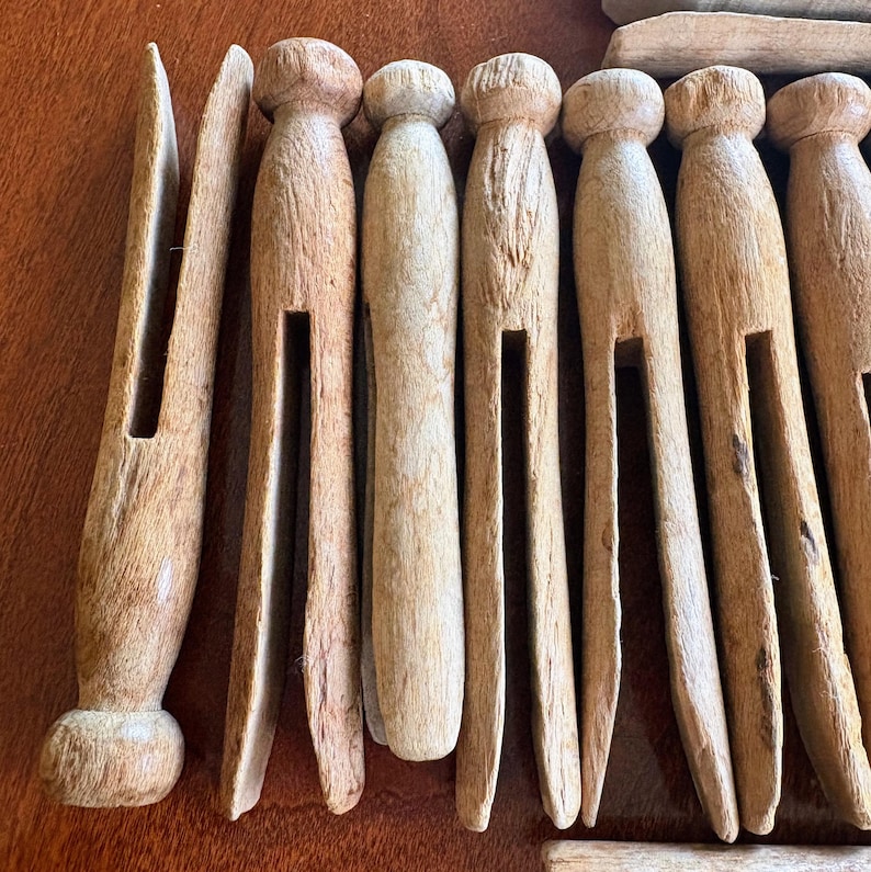 May include: A set of ten wooden clothespins, all in a natural wood color. The clothespins are all in a row, with the top of the clothespins facing the viewer. The clothespins are all in good condition, with no visible cracks or chips.